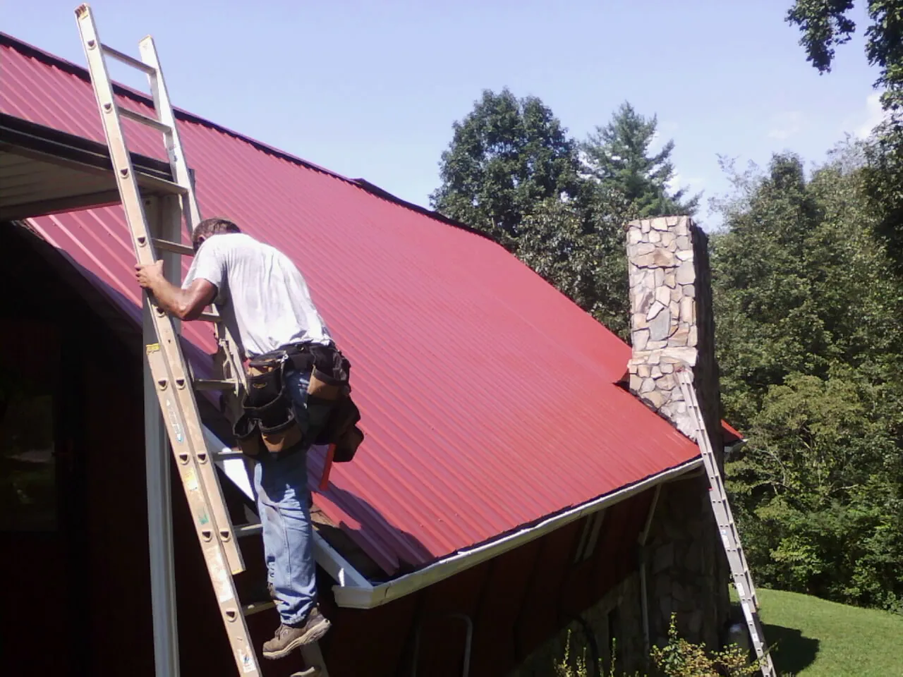 Roofer on a ladder beside a red metal roof and stone chimney — Soffit & Fascia Repair by Roofing Brothers in Laketon
